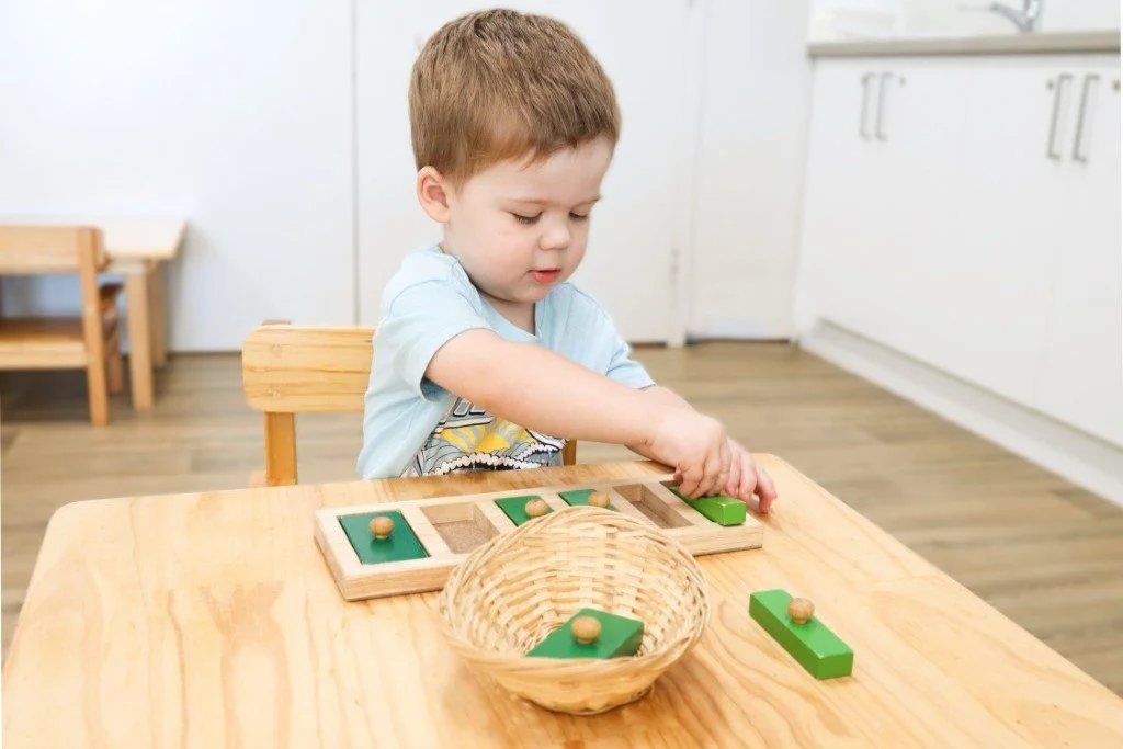 Young boy in a childcare centre, working on a Montessori wooden puzzle pieces on a table in a room with wooden flooring and white cabinets.
