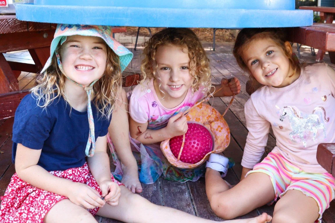 Three young girls sitting under a picnic table outside in Rockingham, smiling at the camera.