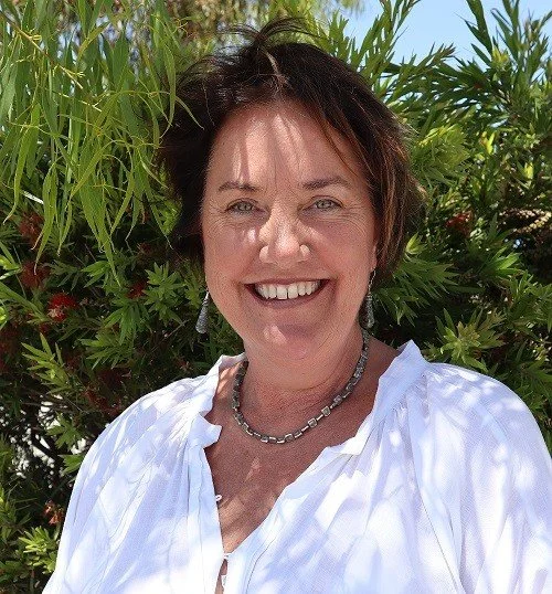 The Principal smiling outdoors surrounded by green plants, wearing a white blouse and jewelry.