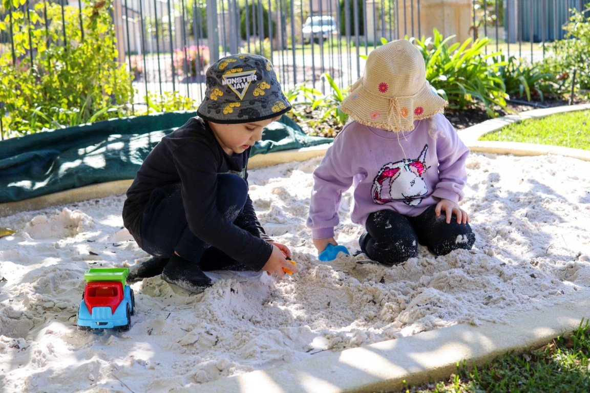 Two children playing in a sandbox outdoors at our Rockingham childcare centre, with one child wearing a black shirt and hat, and the other child wearing a lavender sweatshirt and sunhat, surrounded by greenery.