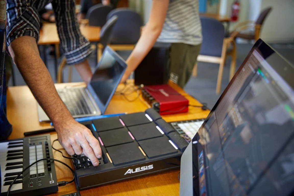 High school student working with electronic music equipment, laptops, and a MIDI controller on a wooden table in a group setting.
