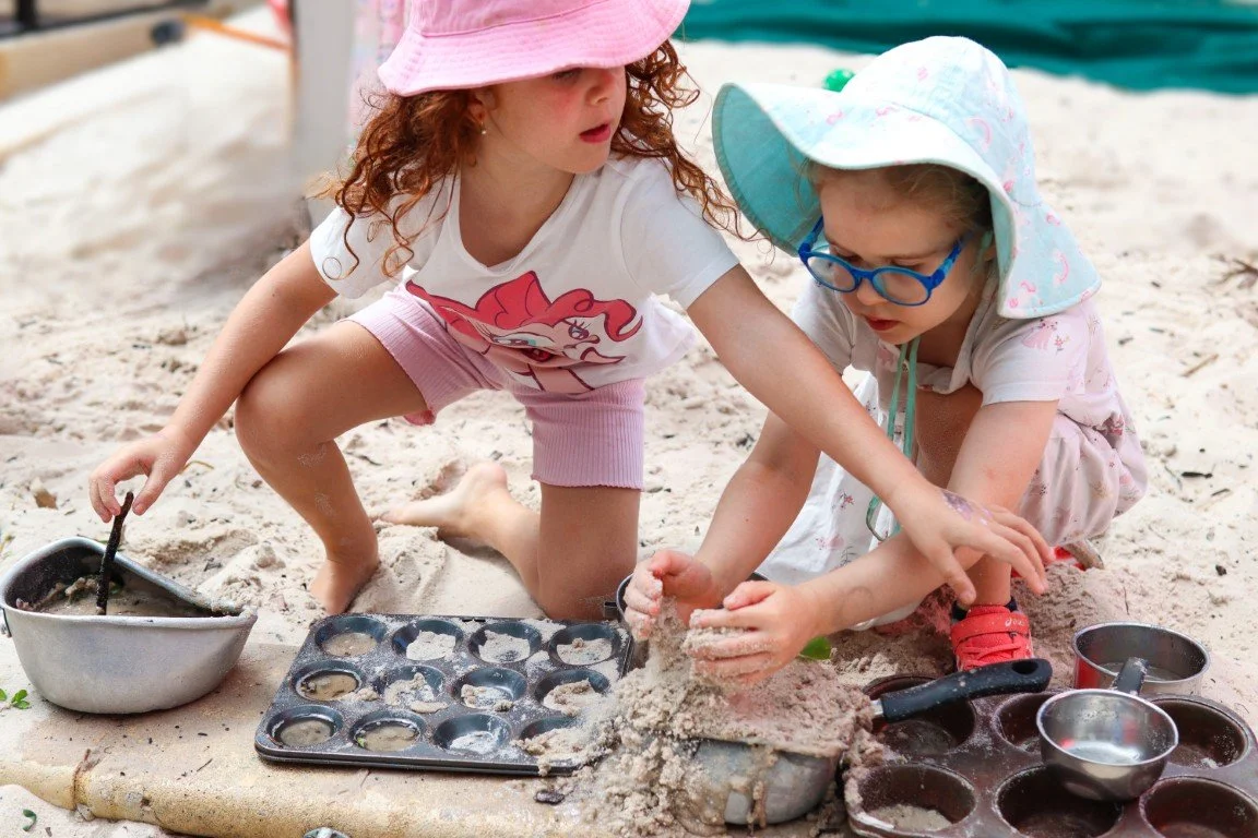 Two young girls playing in the sandpit at the Rockingham Primary School. They are wearing sun hats, one pink and one light blue, and casual summer clothes. Sand toys and baking molds are scattered around them.