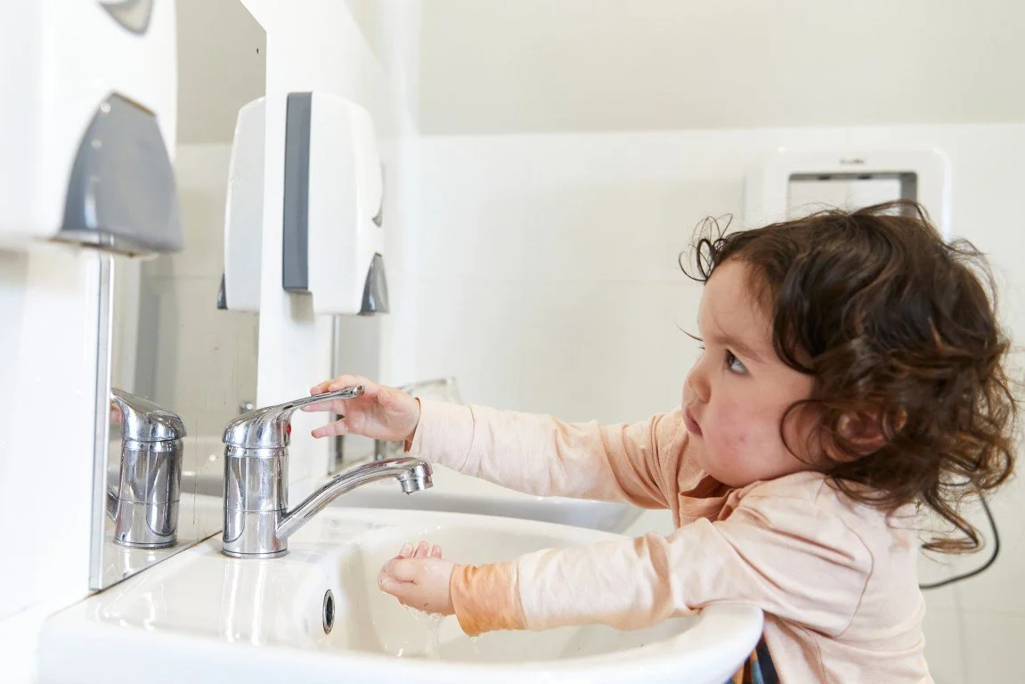 A toddlerl with curly hair washing her hands at a sink in a restroom.