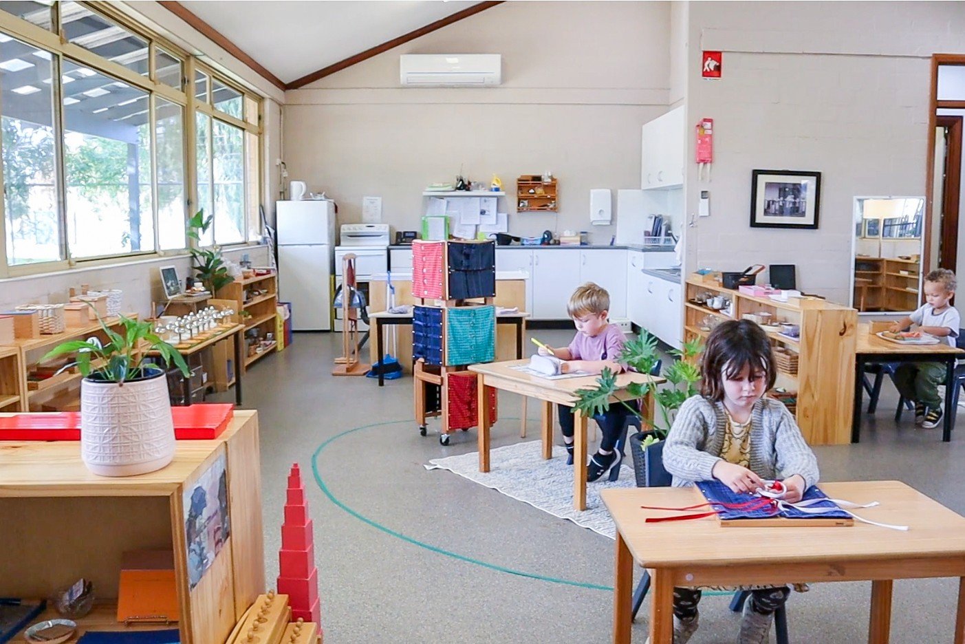 Children working at their desks in a classroom with large windows, plants, and wooden furniture at our daycare in Rockingham.