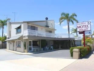 A two-story motel with a white exterior and a balcony on the second floor. There is a sign that says 'Motel' with a phone number. Palm trees and bushes are in the background, and the sky is blue.