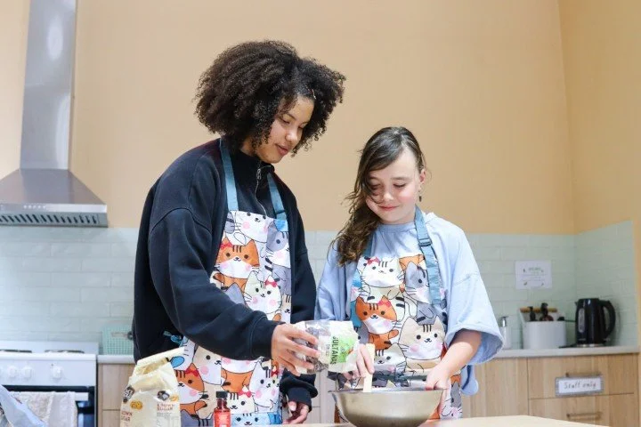 Two primary school girls wearing aprons with cat patterns cooking together in an Upper Primary kitchen