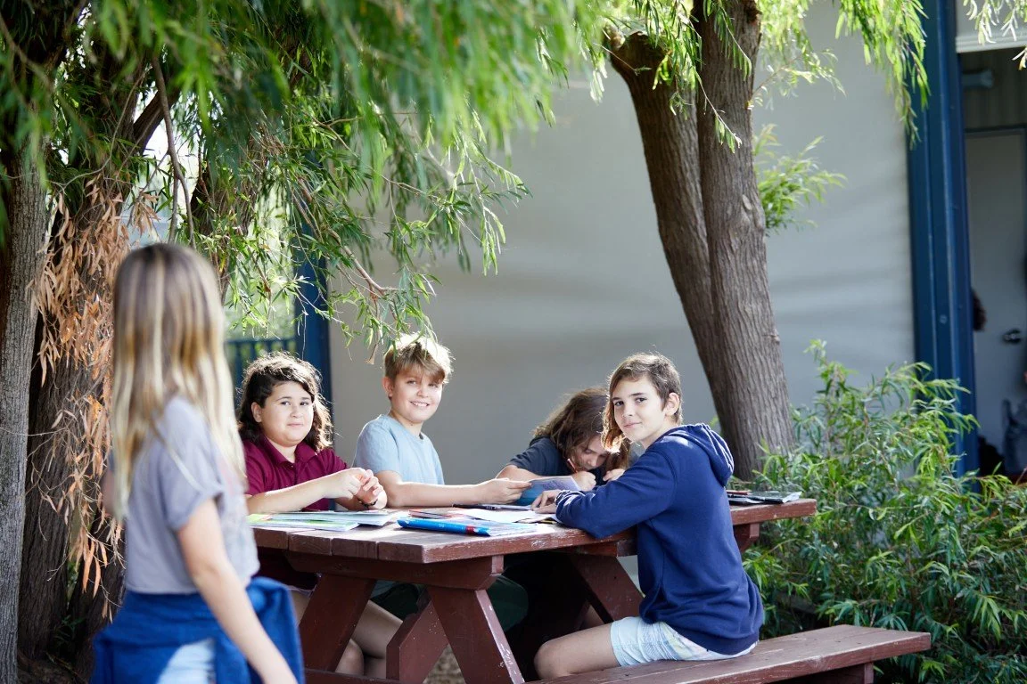 Group of children sitting at outdoor picnic table under trees with books and notebooks at the Rockingham Primary School