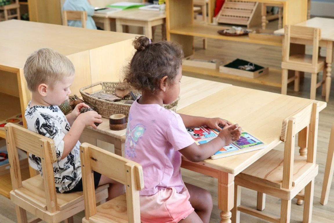 Two children seated at a wooden table in a Montessori Early Learning classroom, one girl reading a colorful book and a boy looking at a small object in his hands, surrounded by wooden furniture and shelves.