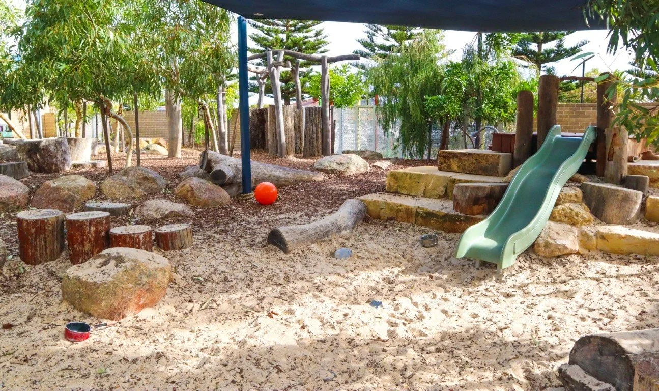 An outdoor playground area at our Port Kennedy Montessori daycare centre with a green slide, wooden logs, rocks, and trees, surrounded by a sandy surface and a blue shade canopy.