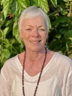 A woman smiling, wearing a white top, a red beaded necklace, and earrings, standing in front of green foliage.