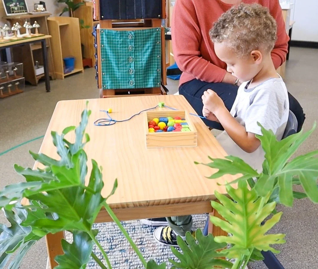 A young boy in Children's House is sitting at a wooden table doing the beading activity, with an adult assisting.