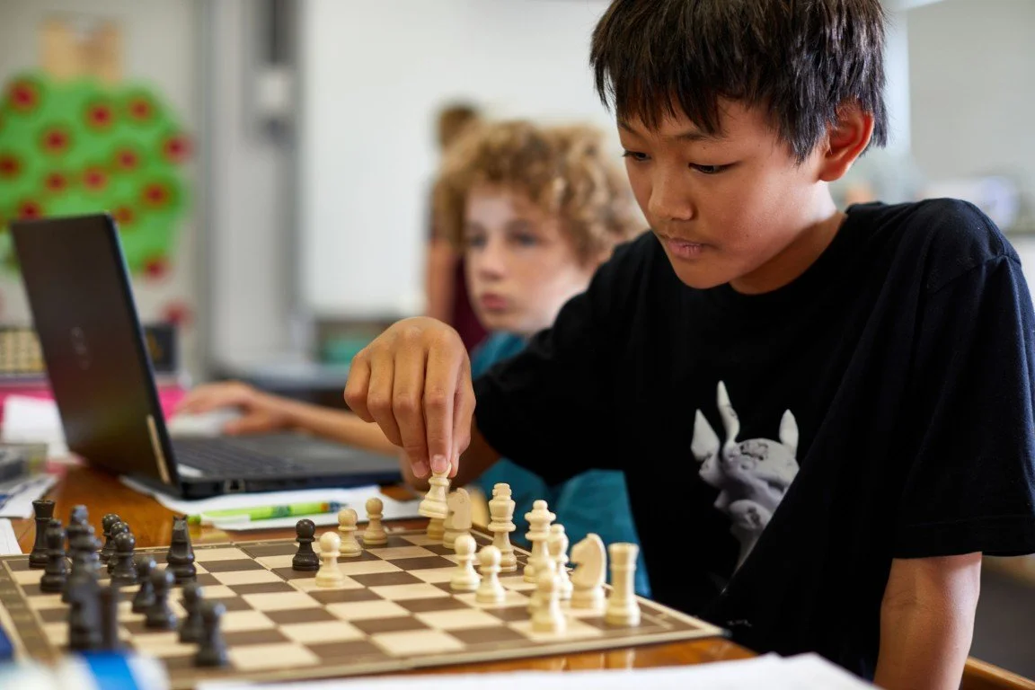 A young boy playing chess, making a move on the chessboard, with a girl and a laptop in the background in a classroom setting.