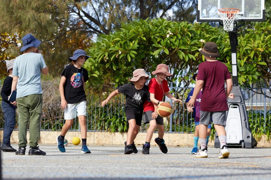 Children playing basketball on an outdoor court at our Rockingham Primary Campus, wearing hats, with some holding a basketball, and a yellow ball on the ground, surrounded by greenery.