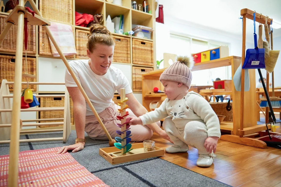 A woman and a young child in Montessori Playgroup. The woman is sitting on the carpet, and the child is squatting next to her, wearing a beige knit hat and a cream-colored outfit. The room has wooden shelves and various toys and storage baskets.