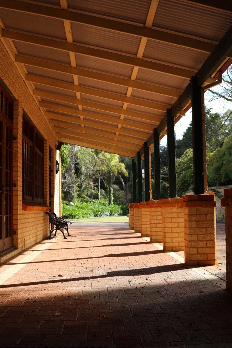 View of a porch with brick columns, a roof, and a bench, overlooking a garden with trees and bushes.