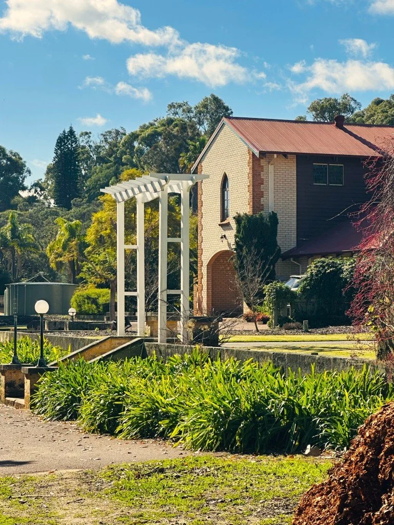 A sunny scene of the venue with a brick facade, arched window, and red roof. In the foreground, there's a garden with lush green plants, a white pergola, and a pathway with decorative light fixtures.