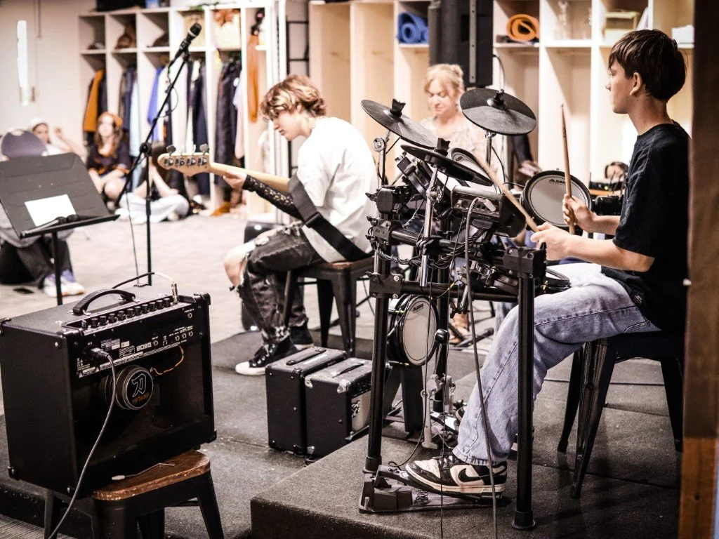 A group of young people playing musical instruments in their high school band, including a girl on electric guitar, a boy on drums, and another person in the background, in a room with open lockers and musical equipment.