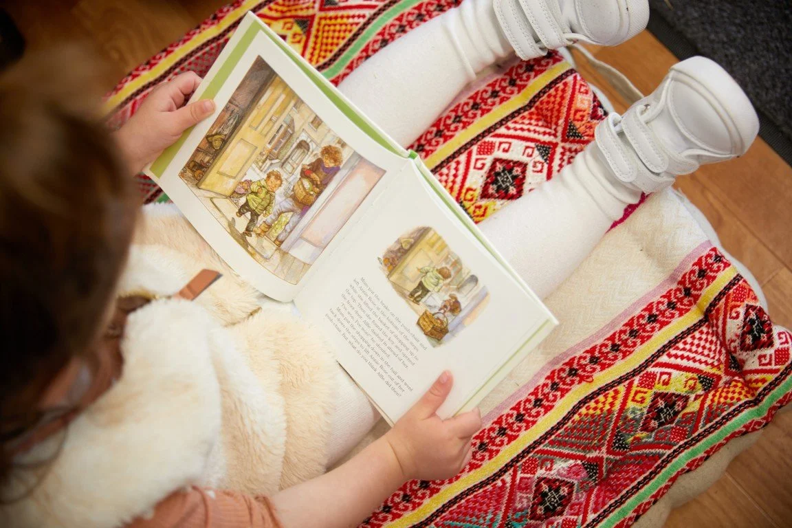 A young girl sitting and reading a children's picture book with colorful illustrations, on a patterned blanket, wearing white boots and white leggings at our Infant Toddler Program.