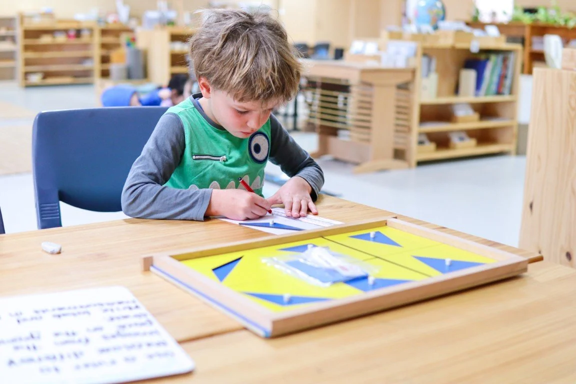 A Lower Primary school student sits at a wooden table in a classroom, writing on a piece of paper with a red pen and a Montessori activity in front of him. The classroom has bookshelves and educational materials in the background.