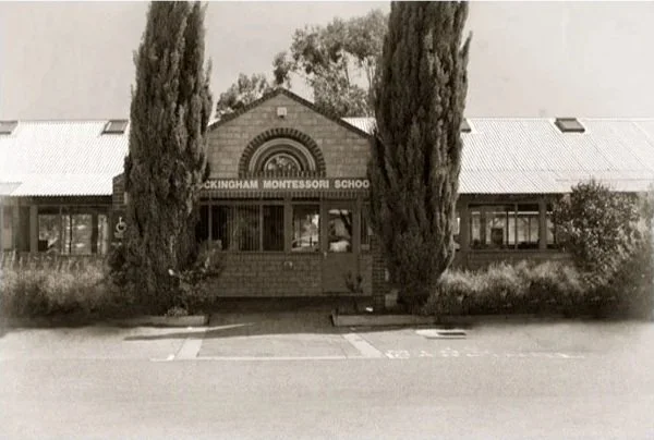 Old photo of a front view of a building labeled 'Rockingham Montessori School' with two tall cypress trees in front and a small parking area.