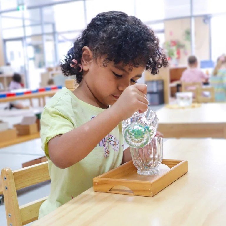 A young child in Kindergarten, wearing a yellow shirt pouring water from a glass pitcher into a clear glass on a wooden tray in a brightly lit classroom.