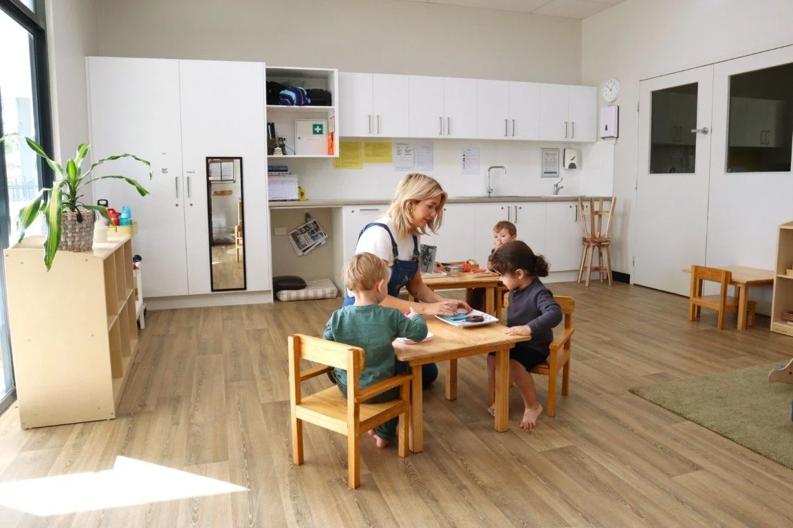 A caregiver in a daycare room with three children at small wooden tables, engaging in an activity. The room has white cabinets, a large window, and a wooden floor.