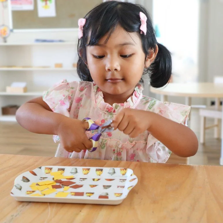 A Montessori childcare student with pink hair clips cutting paper with purple scissors at a table.