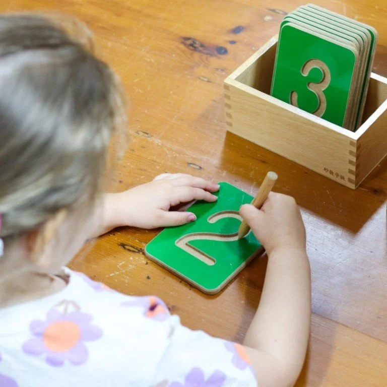 A young child in Children's House engaged in a number tracing activity, using a wooden stylus to trace the number 2 on a green learning board. A wooden box containing flashcards with the number 3 is nearby.
