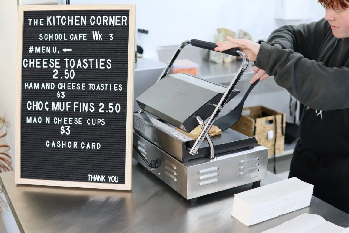 An Adolescent Program student using a sandwich press at a counter with a menu board listing cheese toasts, ham and cheese toasts, chocolate muffins, and mac n cheese cups for sale.
