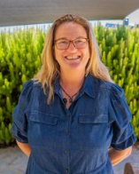 Louise wearing glasses and a denim shirt standing outdoors with green plants in the background, smiling.