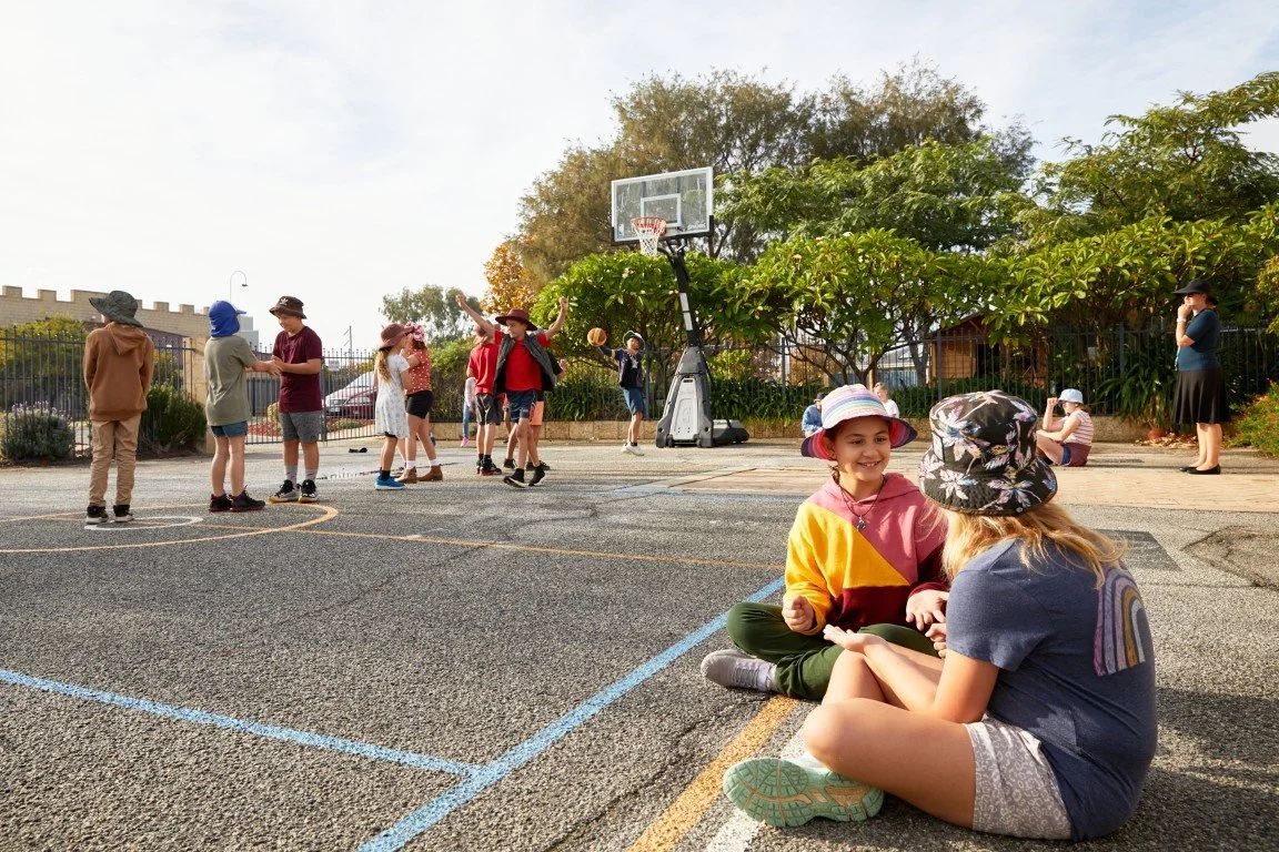 Children playing basketball outdoors while some kids sit on the ground and talk at our Rockingham Primary School.
