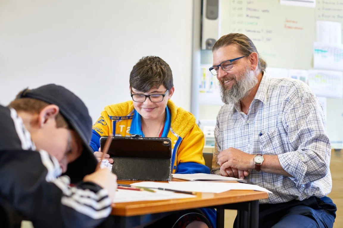 A teacher and two students smiling at a desk in a classroom.