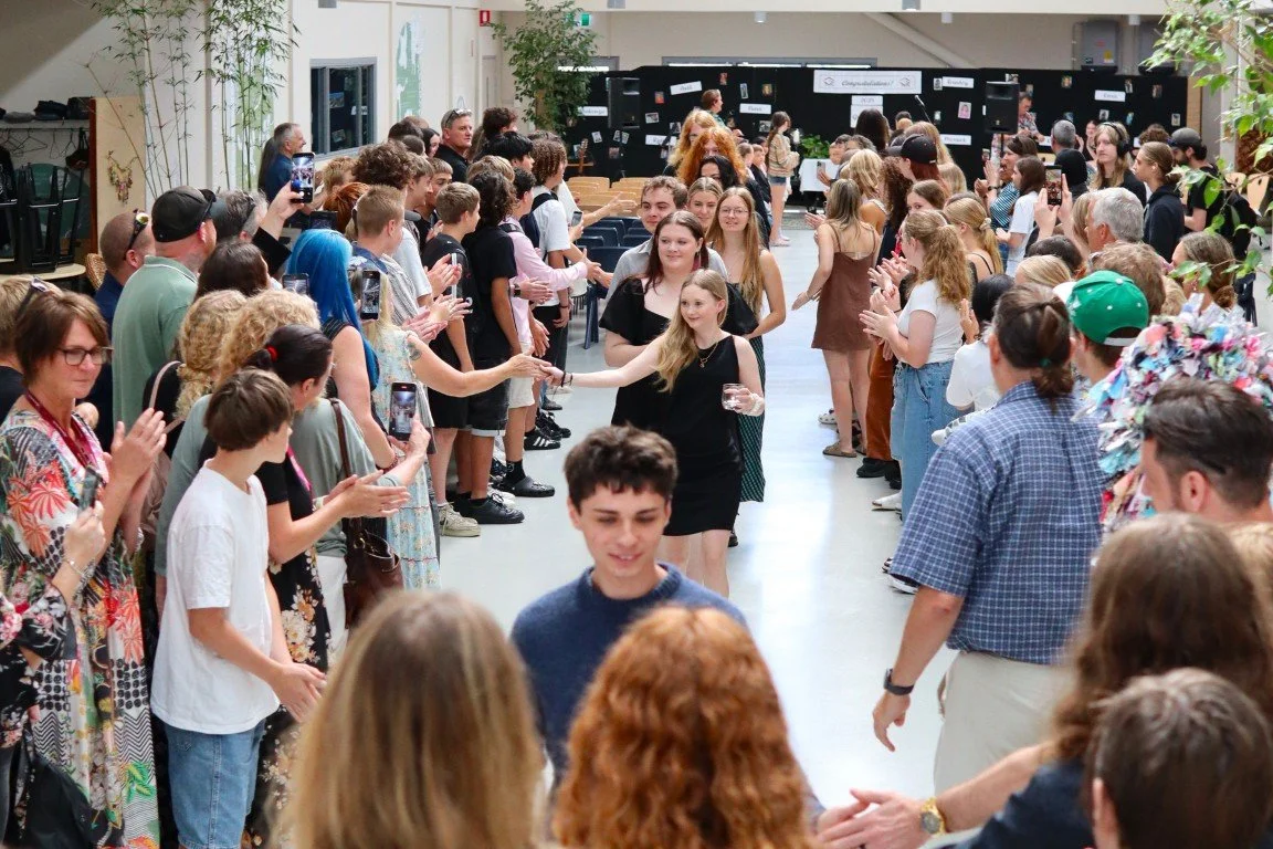 People at a high school graduation event, with individuals lining both sides of a walkway, clapping as a young woman and man walk through.