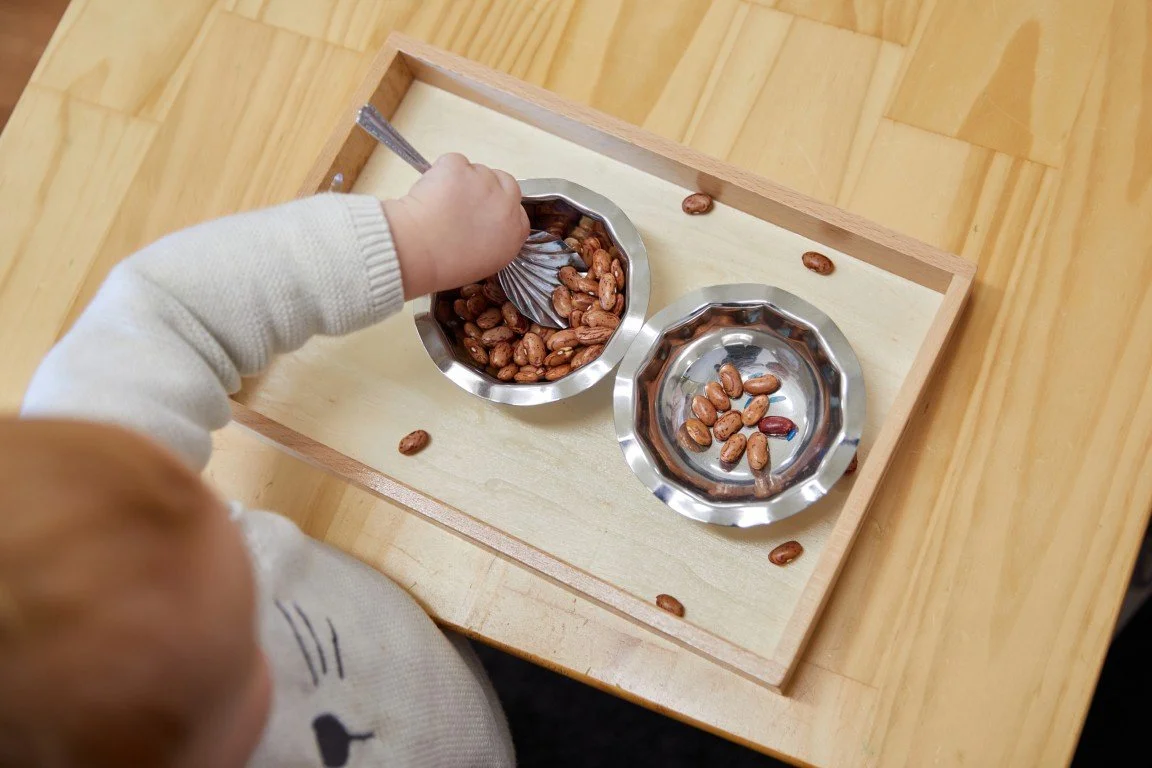 A young child in a sweater is scooping from a metal dish into another empty metal dish on a wooden tray at Montessori Playgroup