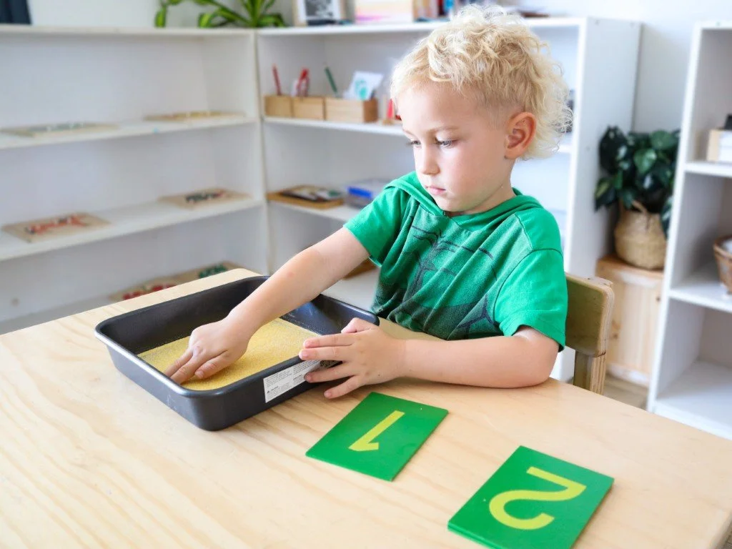 An early learning student with blond curly hair in a green shirt uses his hand to trace the number one in a tray of sand, with two number cards labeled "1" and "2" on a wooden table, in a classroom setting.