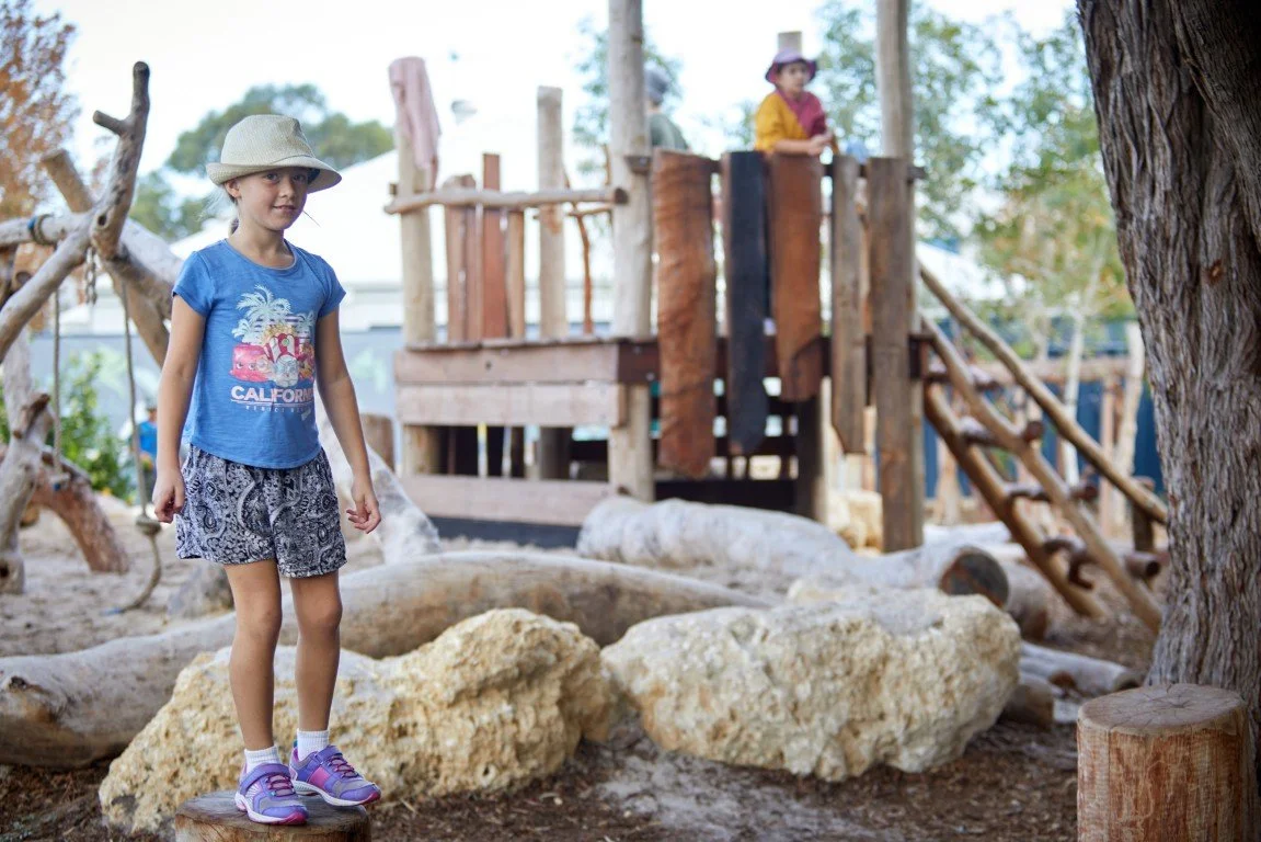 Young girl with a hat standing on a rock in the Rockingham Primary School playground with a wooden structure in the background.