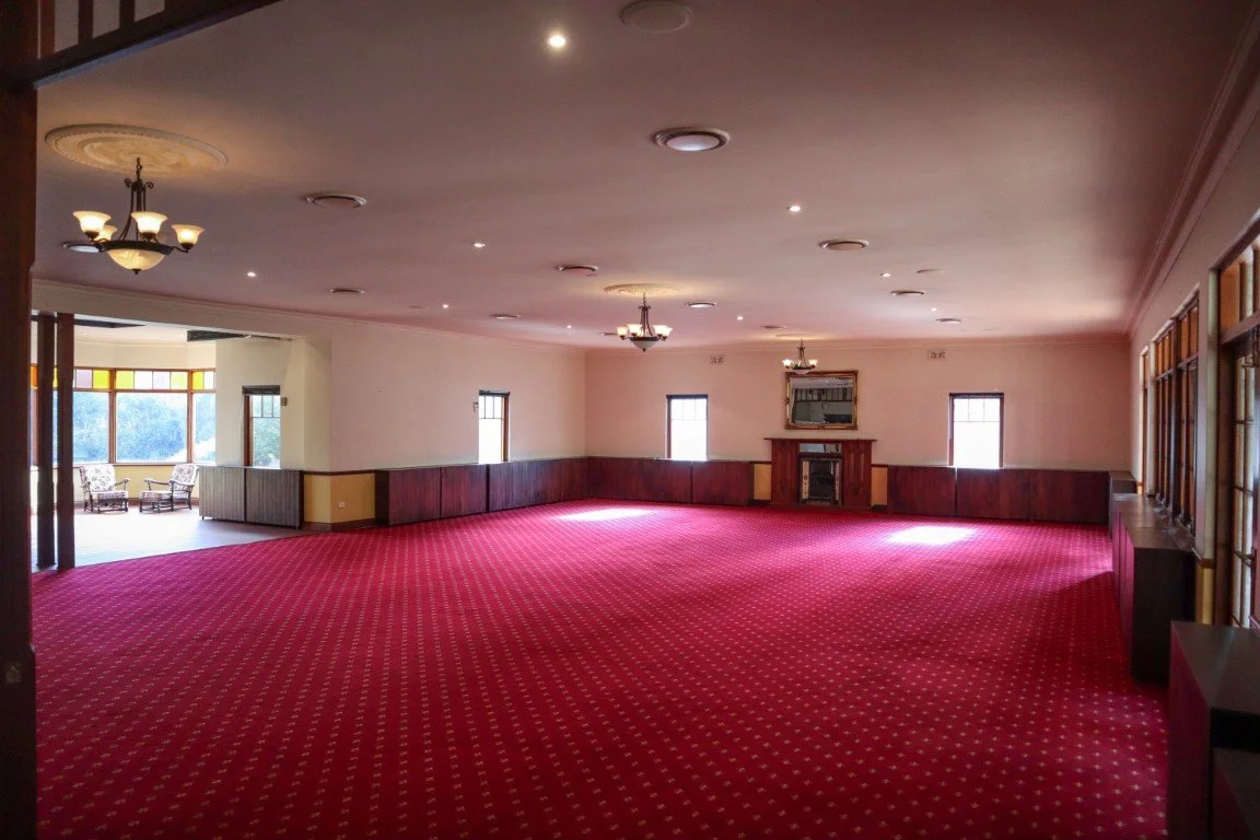 Empty banquet hall with red carpet, wood-paneled walls, small windows, chandelier, and fireplace, with some chairs in an adjacent room.