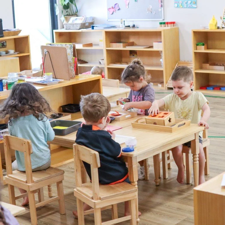 Children sitting around a wooden table engaging in activities in a Montessori Early Learning classroom. The classroom has shelves with educational supplies and artwork on the wall.