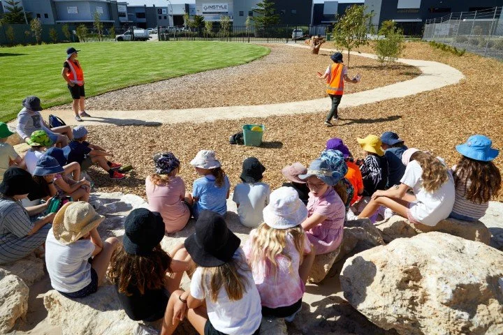 Group of primary school children sitting on rocks in their outdoor playground, watching a person in a safety vest giving a presentation outdoors.