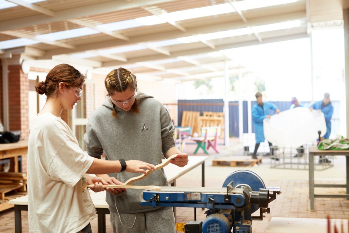 Two students working together on a woodworking project in a workshop, with three people in blue coats in the background holding a large white object.