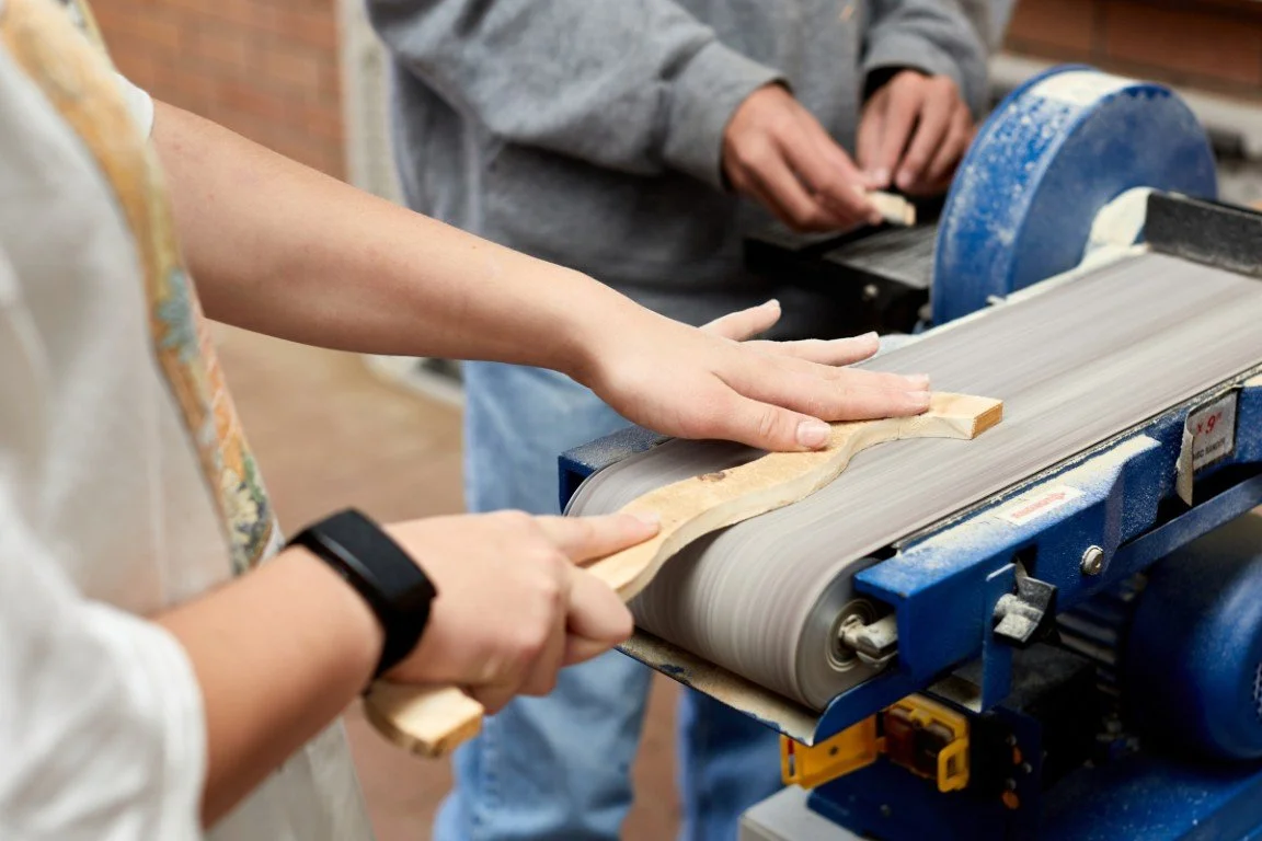 High School students working together in a woodworking shop, using a machine to shape a piece of wood.