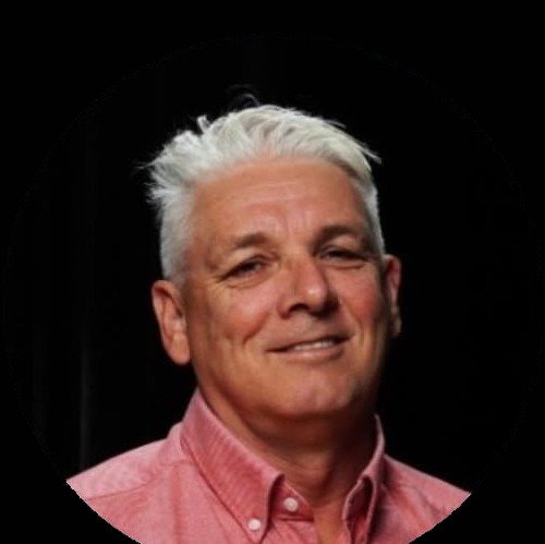 Wayne smiling, wearing a pink button-up shirt, against a black circular background.