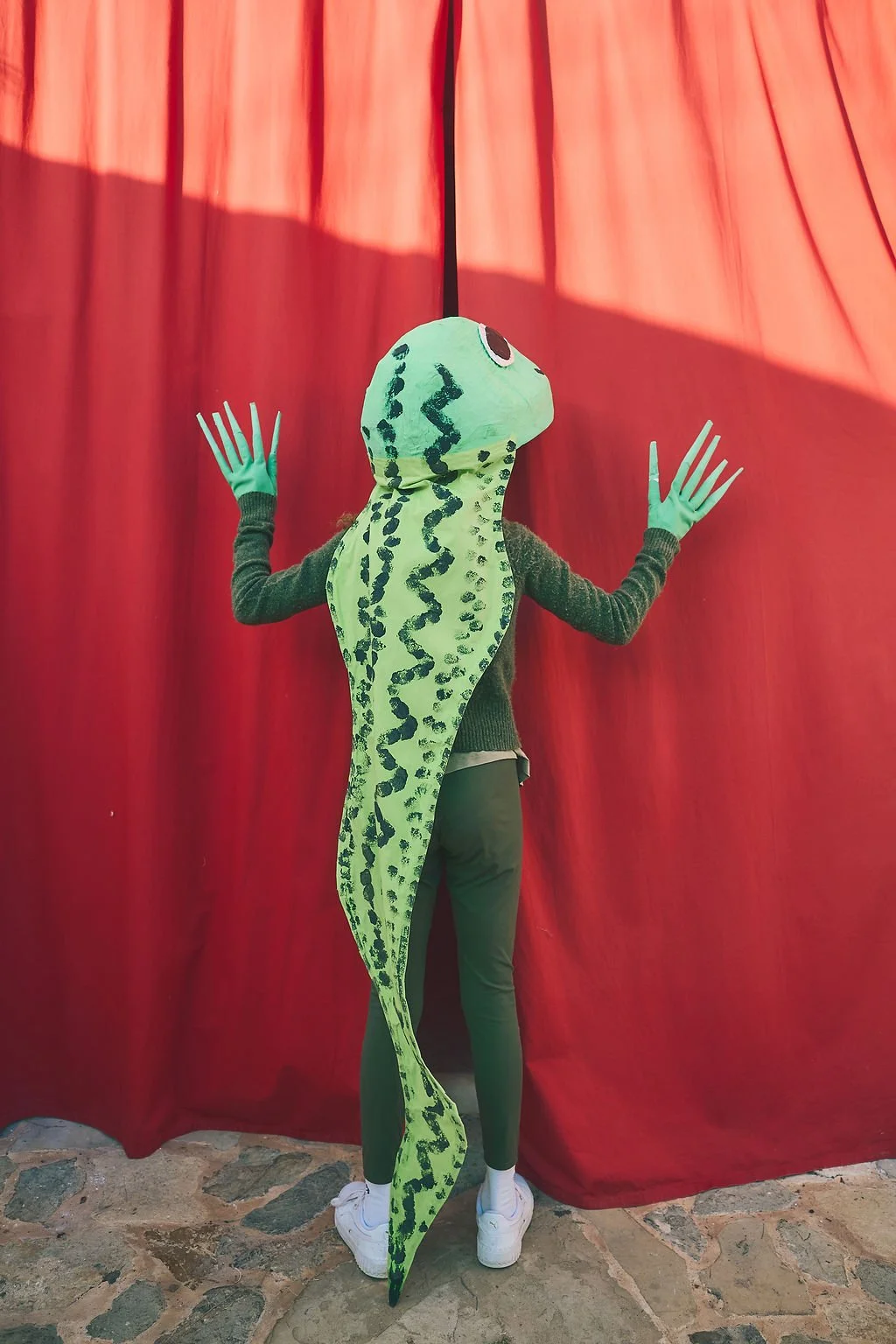 Persona con disfraz de lagarto en un telón rojo, usando guantes verdes y zapatillas blancas.