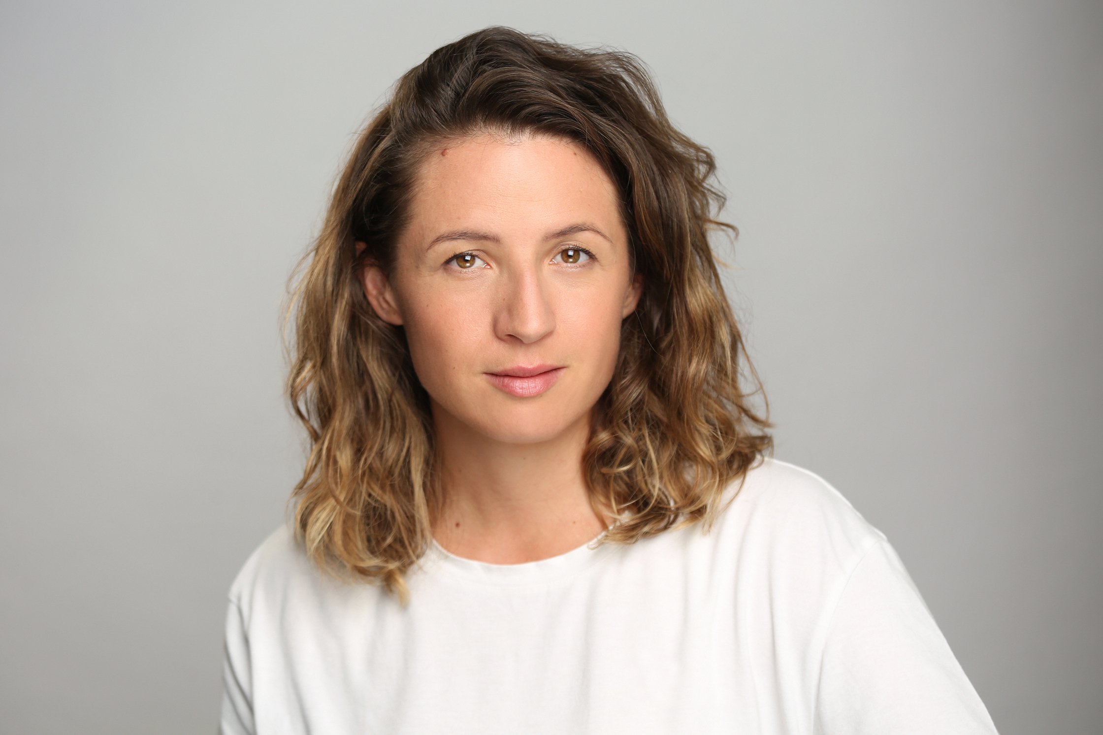 Retrato de una mujer con cabello rizado de longitud media, tez clara y expresión neutral, usando una camiseta blanca.