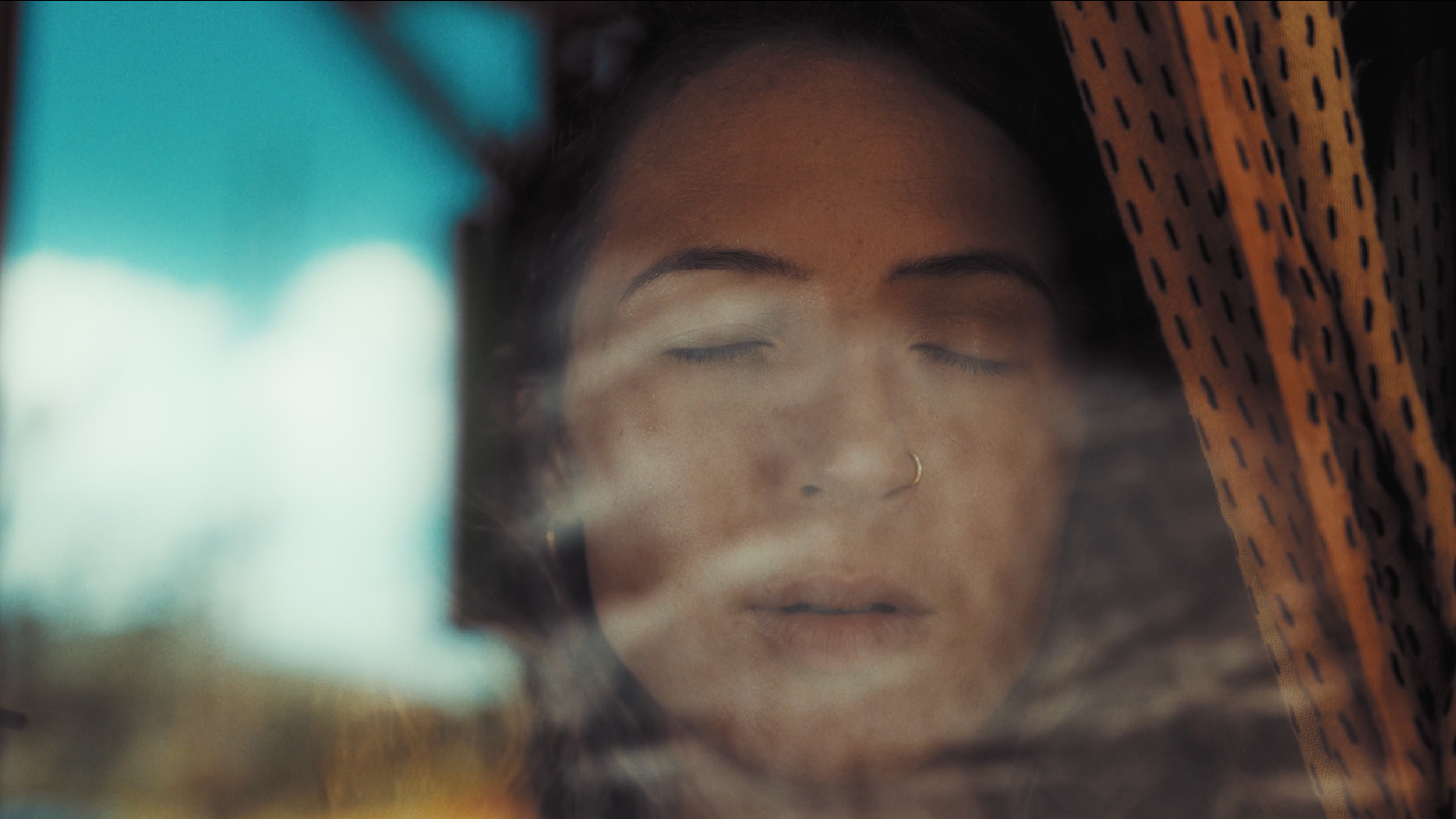 Mujer con los ojos cerrados, vista a través de una ventana con reflejos y cortinas naranjas con negro.