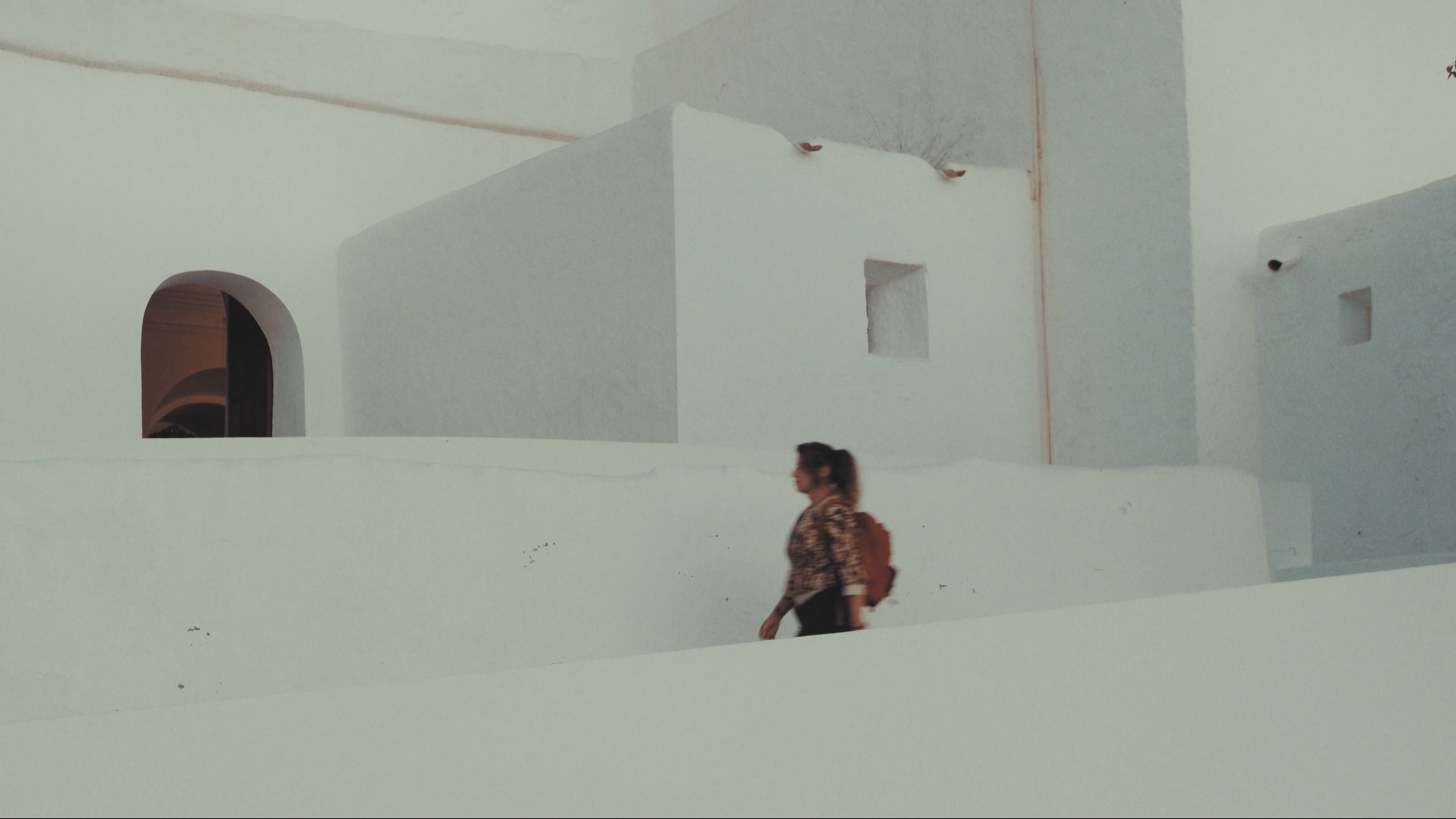 Mujer caminando frente a paredes blancas con ventanas y detalles arquitectónicos en un entorno exterior.