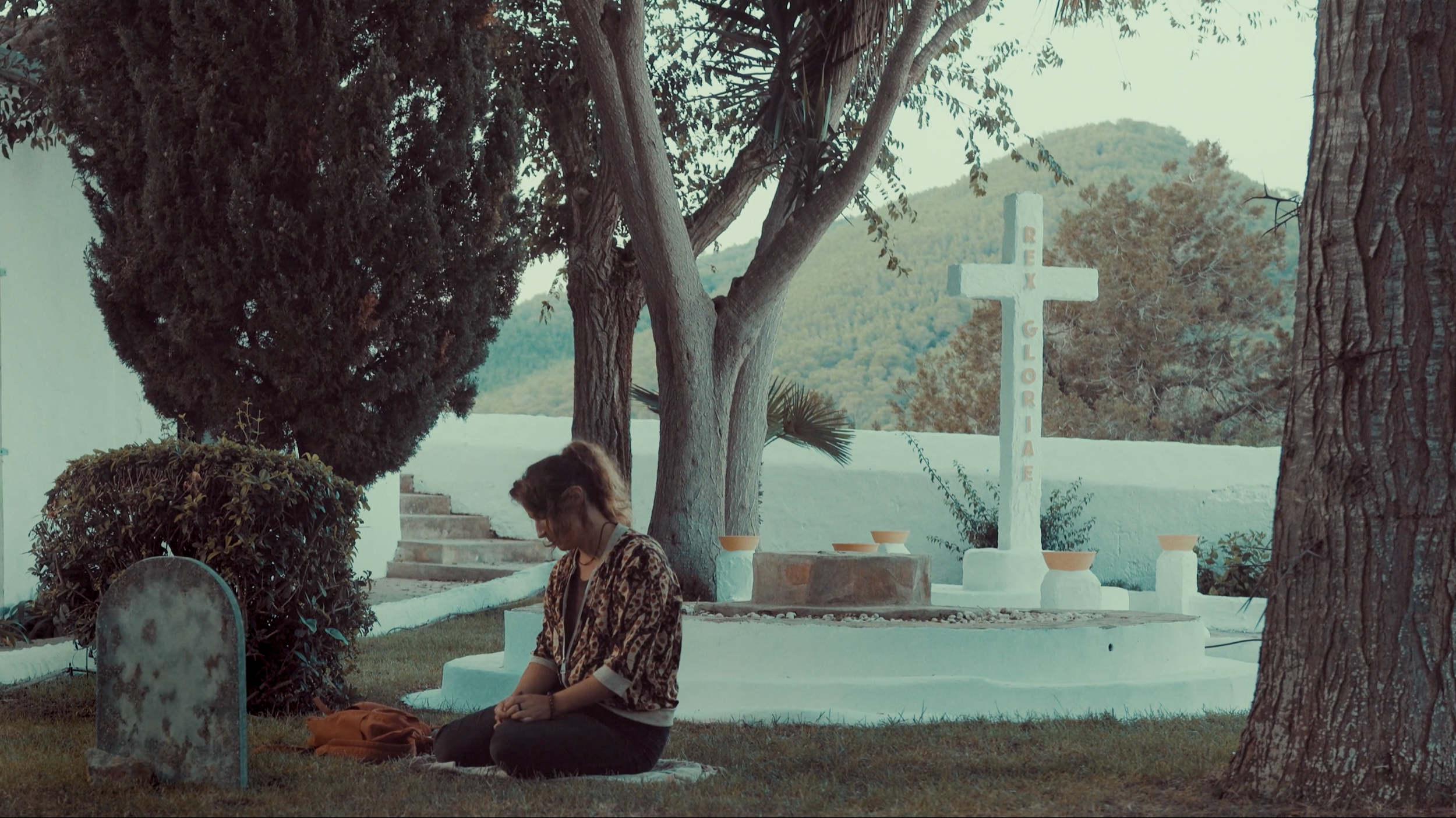 Una mujer en posición de oración en un cementerio con varias tumbas y una cruz grande que dice 'REY GLORIA' en medio de árboles y montañas en el fondo.