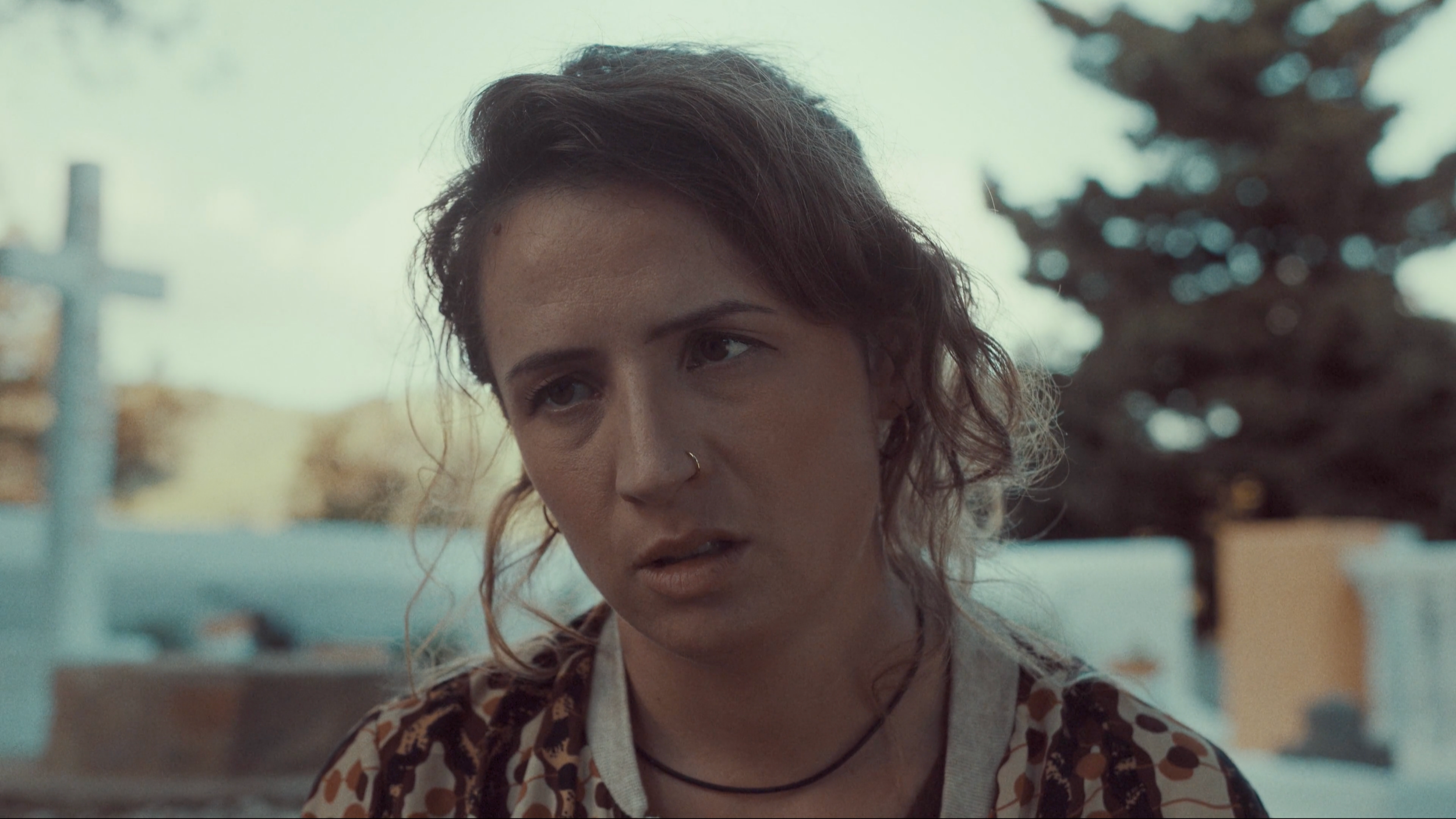 Mujer con expresión de frustración o cansancio, con cabello rizado y bigote, usando camiseta con estampado colorido, en un entorno exterior con árboles y estructura difusa