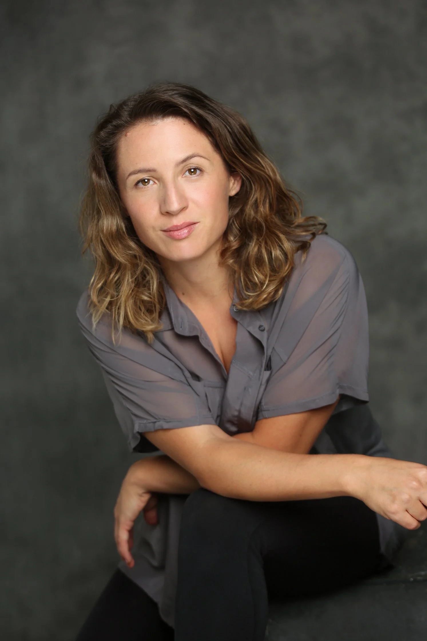 Retrato de una mujer con cabello rizado de color castaño claro, usando una blusa gris y pantalones oscuros, posando en un fondo gris.