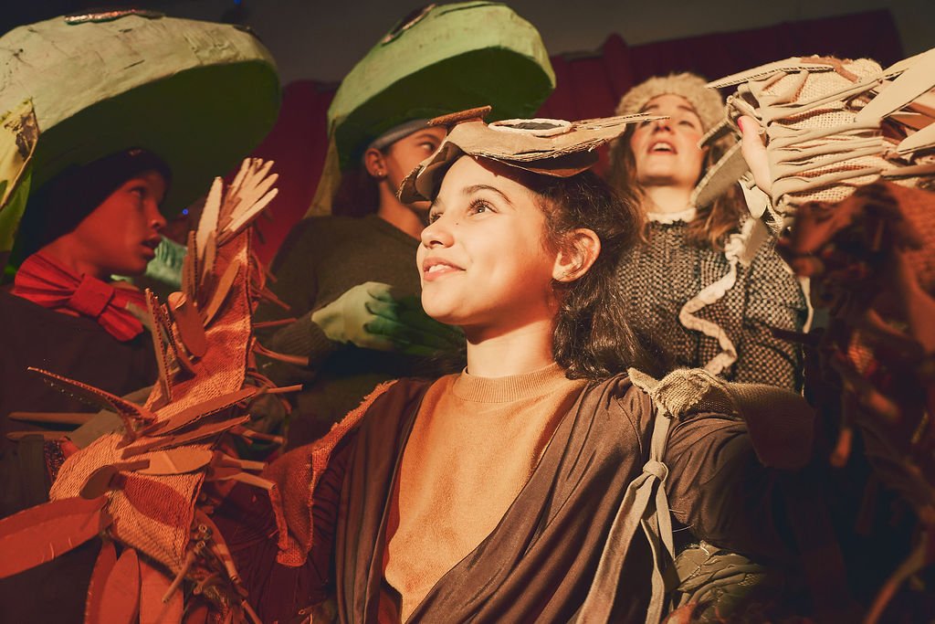 Mujeres participando en una obra de teatro o performance con disfraces de materiales reciclados y elementos naturales.